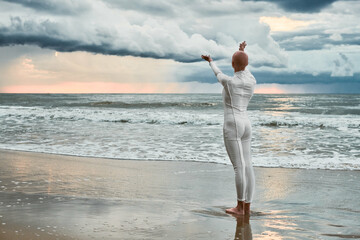 Hairless girl with alopecia in white futuristic suit standing on sea beach stretched out arms to...
