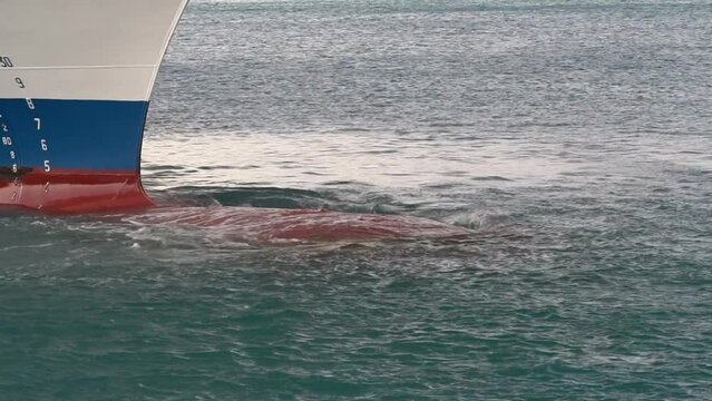 Waves are rolling through red bulbous bow partly covered wit green algae of the merchant vessel with blue and white hull mooring in Bridgetown. On the left are visible draft marks of the ship. 