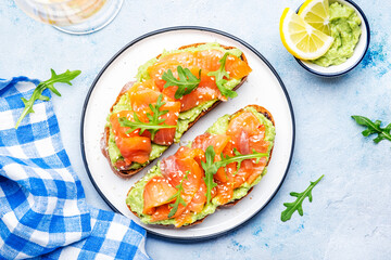 Avocado toast with salmon on rye bread with guacamole sauce, arugula and sesame seeds on plate, blue table background, top view