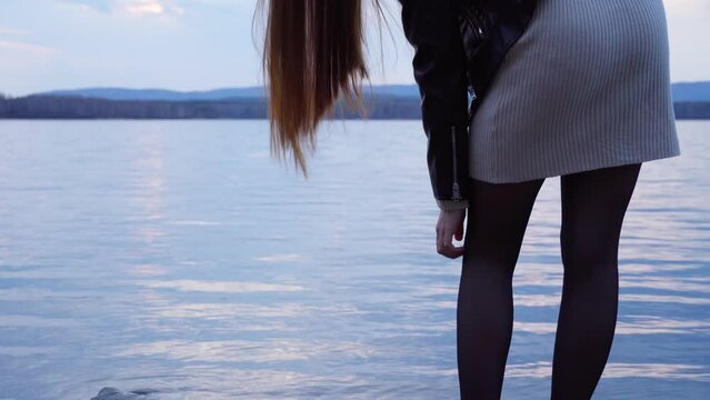 A young woman in black tights and a tight skirt scratches her leg after being bitten by a mosquito. Close-up of the legs of a girl standing on the shore of a lake.