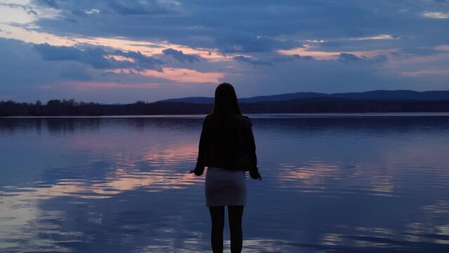 A happy young girl admires the beauty of twilight nature on the shore of the lake after sunset. A mirror image of the sky on the surface of the water.  The harmony of nature and man.