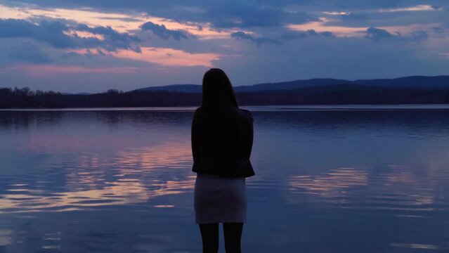 The silhouette of a lonely young girl standing at dusk on the shore of the lake after sunset. The girl admires the beauty of nature. A mirror image of the cloudy sky on the surface of the water.