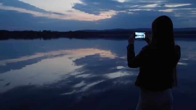 A young girl takes pictures on her phone of the landscape on the shore of the lake after sunset. A mirror image of the cloudy sky on the surface of the water. A woman is happy in nature.