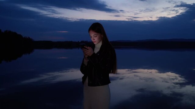 A young girl looks at photos taken on her phone at dusk on the shore of the lake after sunset. A mirror image of the sky on the surface of the water.
