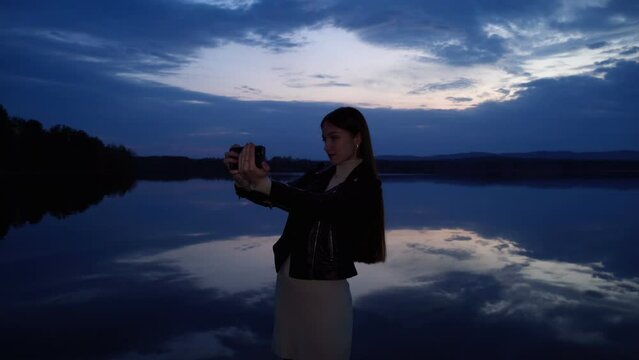 A young girl takes pictures of herself on her phone at dusk on the shore of a lake after sunset. A landscape with a mirror image of a cloudy sky on the water. A girl takes a selfie in nature.