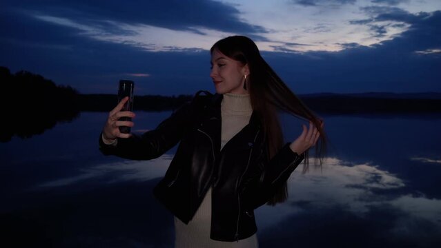 A young girl takes pictures of herself on her phone at dusk on the shore of a lake after sunset. A landscape with a mirror image of a cloudy sky on the water. A girl takes a selfie in nature.