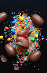 Chocolate eggs and rabbit, sugar colored sprinkles on a black background, top view. Easter...