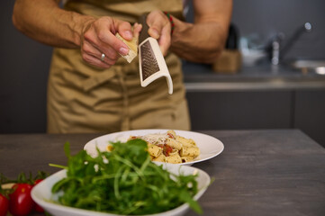 Closeup man cooking dinner at home, preparing Italian pasta with tomato sauce and pouring grated parmesan cheese. A bowl with fresh clean arugula leaves on the foreground