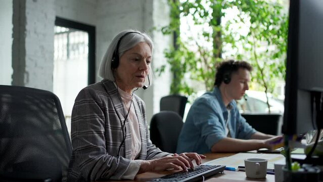 Young corporate worker with headset making phone call. Two colleagues in various age groups in office. Age diverse team concept.