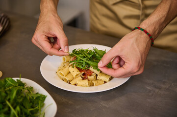 Close-up view from above of the hands of a chef decorating meal with fresh arugula leaves, cooking pasta with tomato sauce according to traditional Italian recipe. Cuisine. Culinary. Epicure