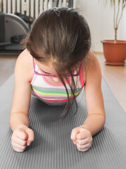 A little caucasian girl in a tracksuit and glasses does a plank exercise, clenching her hands into fists on a gray rug .