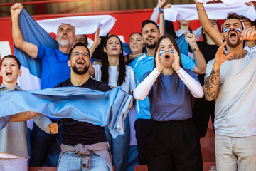 Fototapeta premium Sport fans cheering at the game on stadium. Wearing blue and white colors to support their team. Celebrating with flags and scarfs.