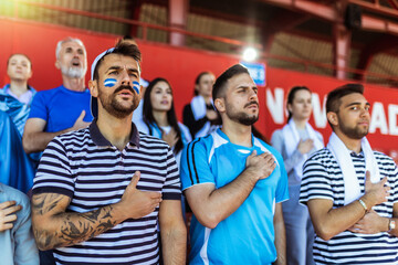 Sport fans singing national anthem of their country at the stadium. Wearing blue and white to support their team.