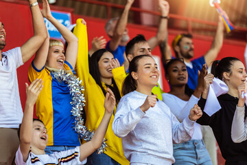 Sport fans cheering at the game on stadium. Wearing yellow and blue colors to support their team. Celebrating with flags and scarfs.
