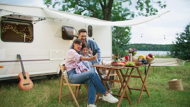 Cheerful Caucasian family having dinner outdoors. Beautiful woman making selfie on phone with her lovely husband and cute daughter. People smiling on camera while standing by caravan car.