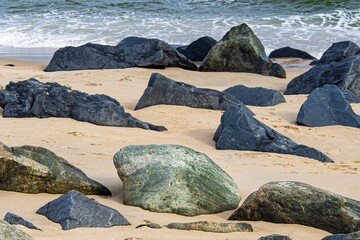 Black, brown and green weathered boulders on a rocky beach, strewn among sand, a hint of waves...