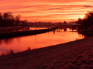 czerwony wschód słońca nad rzeką z sylwetką mostu i ptakami na latarniach - obrazy, fototapety, plakaty
