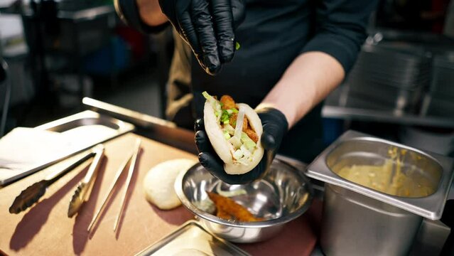 close up chef in black gloves puts different ingredients into a bun with tongs