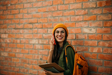 Confident female student standing against a brick wall on campus, holding a stack of books and using a phone