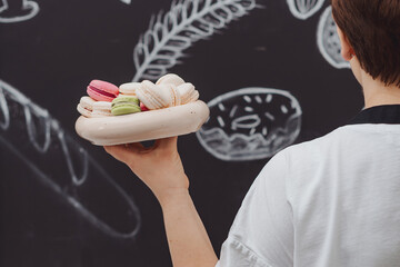The pastry chef is holding a plate with fresh colored macaroons in her hand on black kitchen background.