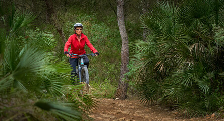 active senior woman riding her electric mountain bike on a djungle trail between palm trees near Xabia, Costa Blanca, Spain