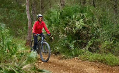 Fototapeta premium active senior woman riding her electric mountain bike on a djungle trail between palm trees near Xabia, Costa Blanca, Spain