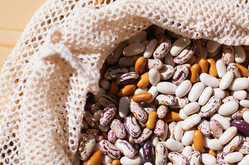 Close up multi-colored beans spilling out from an open, natural fiber mesh eco bag onto wooden...