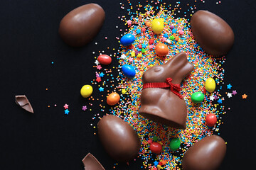 Chocolate eggs and rabbit, sugar colored sprinkles on a black background, top view. Easter...
