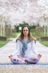 A young woman sits in the lotus position and meditates against the background of a spring blossoming Sakura alley. The path is covered with petals. Concept of mental and physical health and balance.