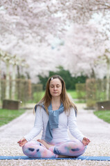A young woman sits in the lotus position with her eyes closed and meditates against the background of a spring blossoming sakura alley. Concept of mental and physical health and balance in life. 