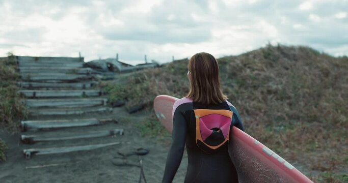 Back, woman and surf at beach, walk up stairs and ready for fitness with wet suit, extreme sport and surfing in nature. Steps, exercise and surfer at ocean with professional or hobby and challenge
