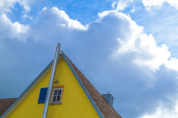 Giebelseite gelbes Haus mit blauen Fensterladen und Edelstahlkamin vor blauem Himmel mit...