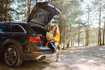 Happy woman searching location on map sitting on trunk of hatchback car outdoors. Leisure, road trip, travel and people concept - obrazy, fototapety, plakaty