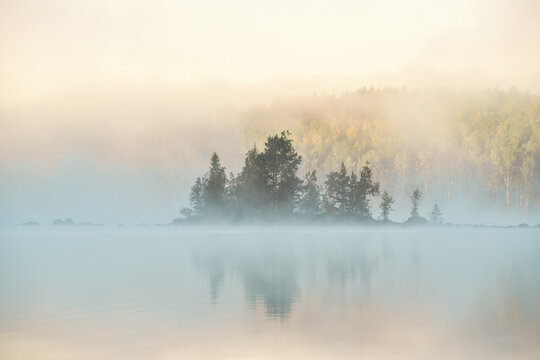 Sunrise over a tranquil lake with a small island, shrouded in mist