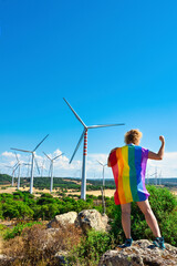 Adult woman on her back with a rainbow flag as a cape, on a rock bending her arm in a landscape with wind turbines.