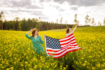 Two young women with american flag on blooming meadow. Summer landscape.  4th of July. Independence...