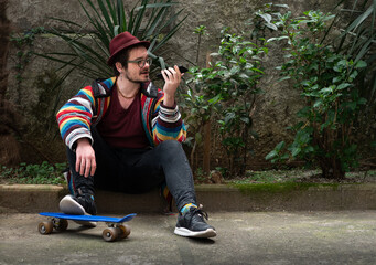 Fashionable young man sitting on skateboard, holding smartphone in garden setting. 