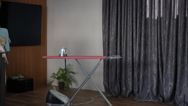 A pregnant woman takes her stack of baby clothes from the table and places them on the ironing board in the living room. The woman waves her head dejectedly and begins to iron the baby clothes with a