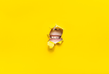 A child's mouth appears in a torn hole of yellow paper. Concept of grin and milk teeth.