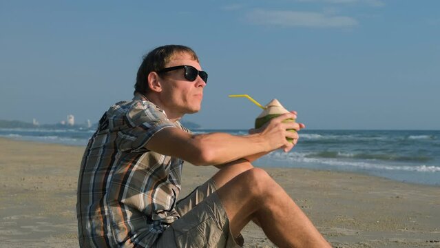 Male in sunglasses drinking refreshing coconut water while sitting on the beach. Travel and vaction concept
