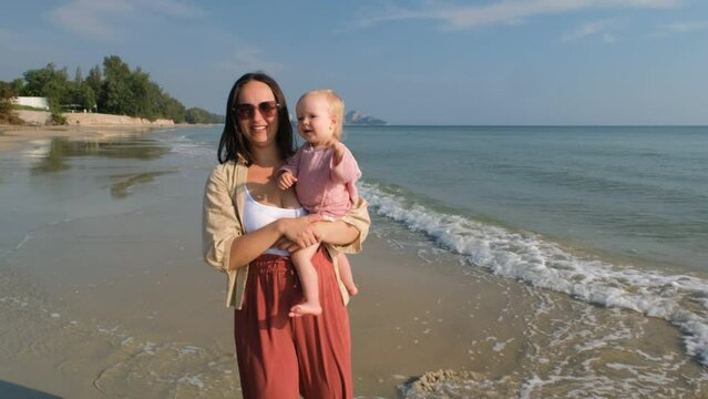 Cheerful young mother with cute baby girl walking along sea shore, slow motion. Family vacation concept