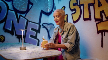Fashion senior drinking cocktail in graffiti cafe. Joyful elderly lady resting