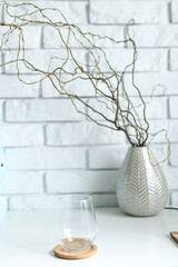 An empty transparent glass stands on a white table top, in the background a decoration of dry twigs and white brick