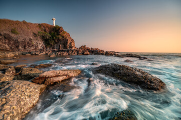 The sun sets behind a solitary lighthouse perched atop a cliff surrounded by a rugged, rocky...