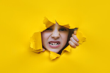 A little girl looks through a torn hole in yellow paper. The concept of anger, fear, fright and...
