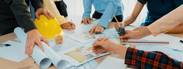 Fototapeta premium Professional architect cooperate with engineer discussing the use of green design in eco house project on table with blueprint and architectural equipment scatter around. Closeup. Delineation.