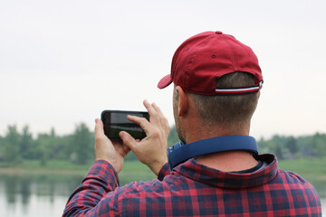 man takes pictures of nature on his smartphone while walking along the riverbank. guy in the plaid shirt and baseball cap, the blogger.