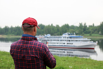man uses a smartphone while standing on the riverbank in summer. he takes pictures of the ship, chats, writes message.