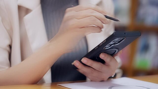 A woman is using a cell phone with a stylus. She is sitting at a desk with a stack of books in the background