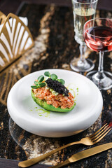 Delicious avocado and raw salmon fish tartare with black caviar on plate on marble table with wine...